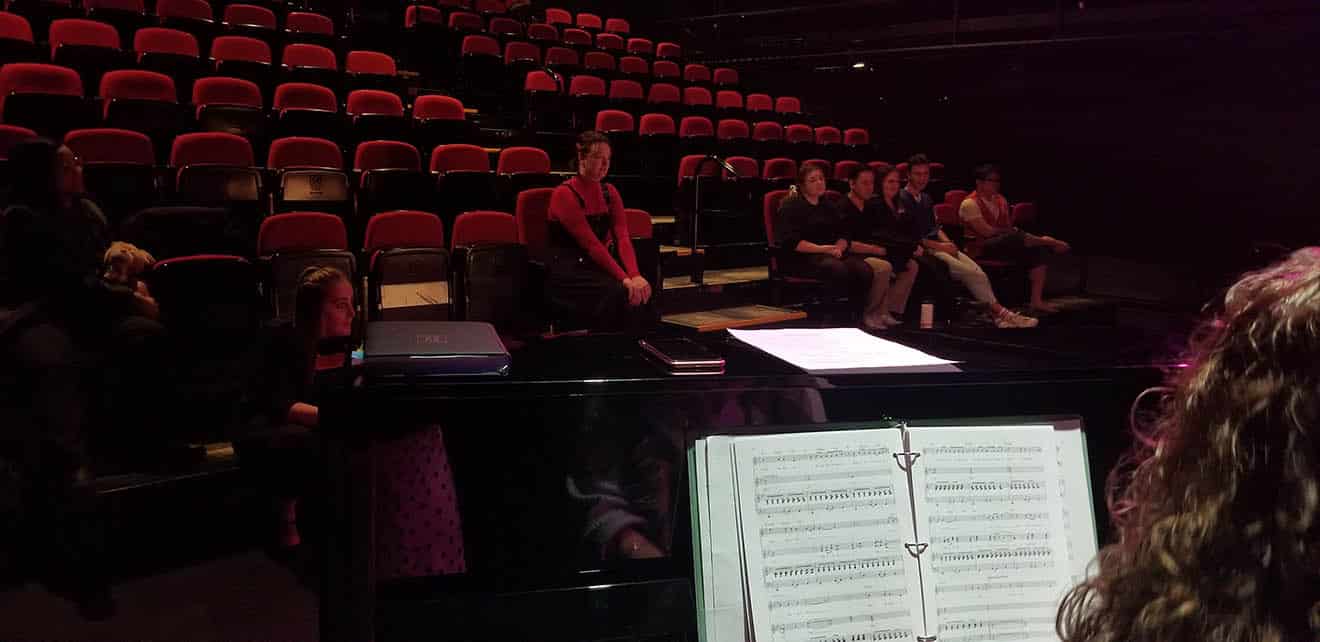 Students in theatre seats looking towards stage. Photo is taken from behind a person at a piano.